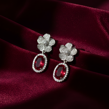 Pair of earrings with red gemstones and diamond-like elements on a burgundy fabric background