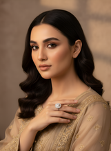 Woman wearing a beige embroidered outfit with a ring on her finger against a neutral background