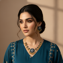 Woman wearing a teal traditional outfit with gold embroidery and jewelry against a beige background