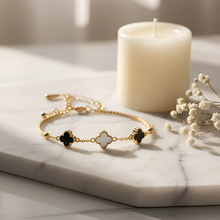Gold bracelet with black and white clover charms on a marble surface with a candle and flowers.